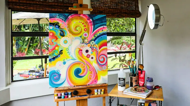 An artist's easel positioned by a window, featuring a colorful painting. A black-armed magnifying lamp provides additional light, set against contrasting grey walls.