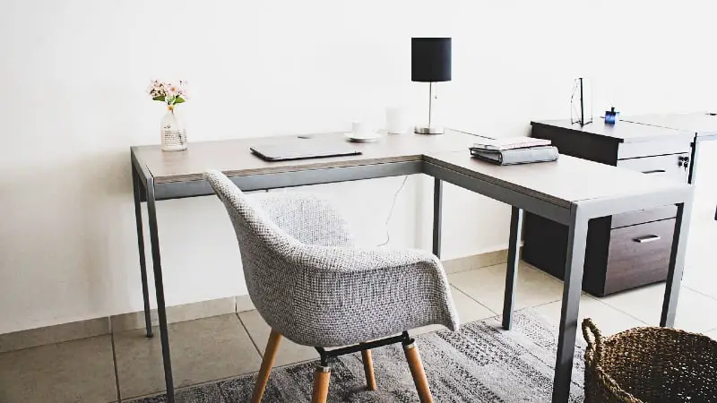 An L-shaped desk with a grey tabletop, featuring flowers and a lamp with a black shade.