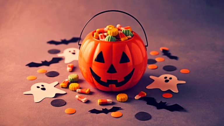 A flat lay photo of retro Halloween items, including a classic plastic pumpkin candy bucket, friendly ghost decorations, and a scattering of candy corn on a dark surface.