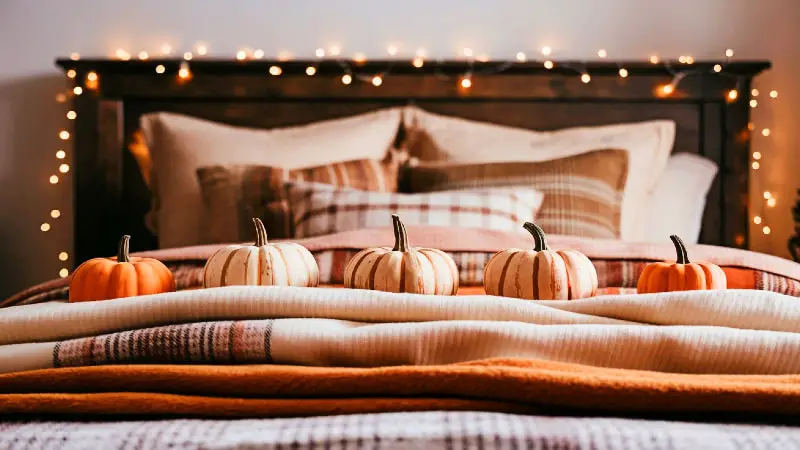 A cozy fall bedroom scene with a bed decorated in orange and cream cotton and flannel blankets. Three small decorative pumpkins and warm fairy lights are arranged on the wooden headboard.